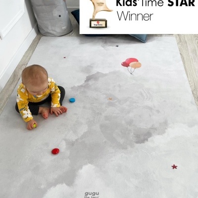 Bebé sentado num tapete cinza claro com balões e estrelas, perto de uma cesta para brinquedos cinza.