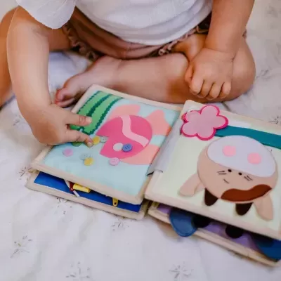 Livro de pano com figuras de feltro coloridas e botões, bebé a tocar.