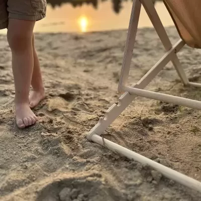 Cadeira de madeira clara com tecido bege na areia junto a lago ao pôr do sol