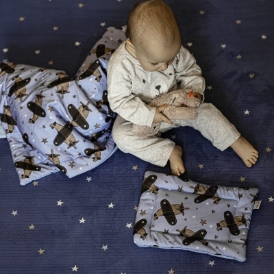 Bebé com pijama cinza segurando peluche rosado sobre tapete azul com padrão de estrelas, com manta e almofada azuis com aviões