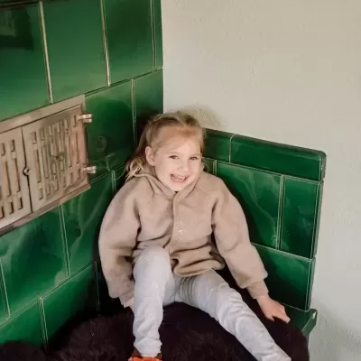 Menina sentada numa lareira de azulejos verdes, roupa bege e cinza, pantufas laranja com raposa.