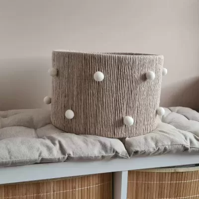 Cesto redondo bege texturizado com bolinhas brancas sobre almofada numa estante branca com cestos de vime.