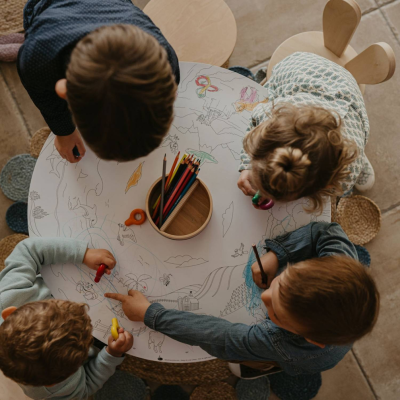 Crianças a colorir numa mesa de atividades branca com desenhos a preto e lápis de cor no centro