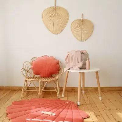 Tapete e almofada em forma de concha cor-de-rosa, cadeirão de rattan, mesa branca com toalha e recipiente, chão de madeira clara.