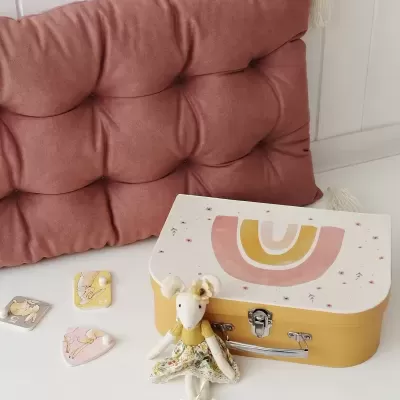 Mala de criança amarelo e branco com arco-íris, boneco de rato vestido, almofada rosa e ímanes coloridos sobre mesa