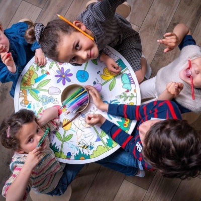 Mesa redonda com desenhos coloridos e cinco crianças a desenhar com lápis de cor numa sala com chão de madeira.