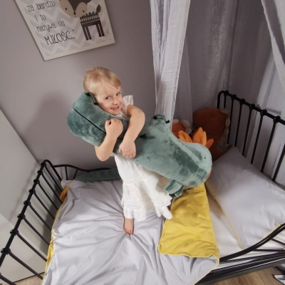 Criança com peluche verde numa cama com estrutura preta e roupa de cama cinza e amarela