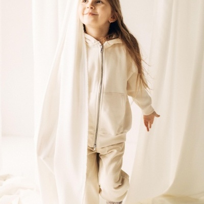 Criança com casaco bege claro e calças bege junto a cortinas brancas