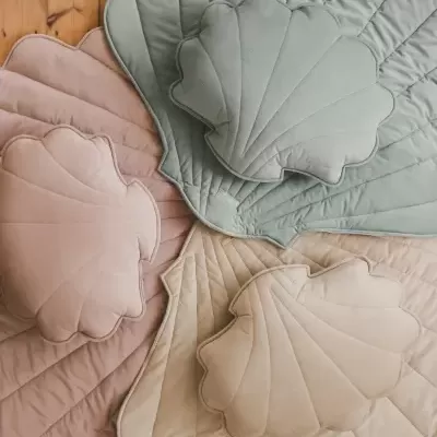 Conjuntos de almofadas e mantas com formato de concha em verde, rosa e bege sobre pavimento de madeira