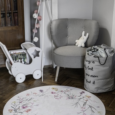 Canto de quarto com poltrona cinza, candeeiro de pé branco, carrinho de madeira branco e tapete floral redondo.