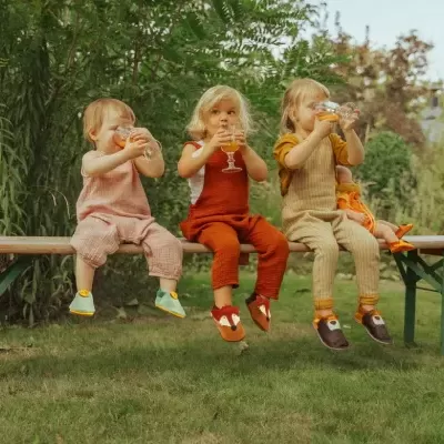 Três crianças vestidas com roupas coloridas sentadas num banco verde bebendo de copos