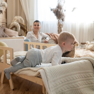 Berço de madeira clara com grades azuis e manta bege com criança e mulher ao fundo em quarto luminoso