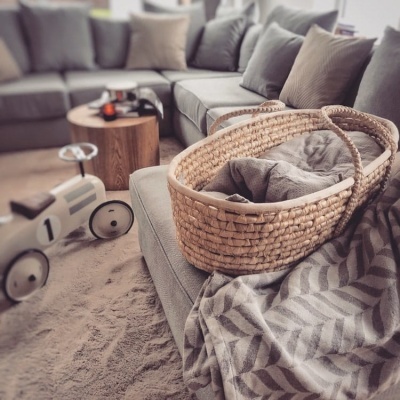 Sala com sofá cinzento, cesto de verga, manta cinzenta e carrinho de brinquedo branco