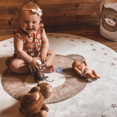 Bebé sentada num tapete redondo com padrão de urso, com três brinquedos ao seu redor.