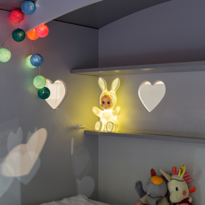 Candeeiro de bebé em forma de coelho com luz quente numa prateleira branca com guirlanda de luzes coloridas e peluches.
