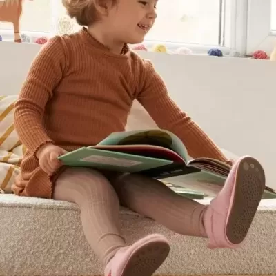 Menina sentada num sofá a ler um livro, vestindo vestido castanho e leggings cor bege.