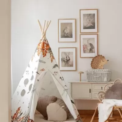 Tenda de brincar tipo tipi com padrão de folhas e pássaros, almofadas, cómoda branca e quadros de coelhos num quarto infantil