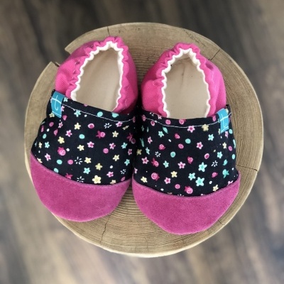 Sapatos de bebé em tecido rosa e preto com padrão floral sobre um tronco de madeira.