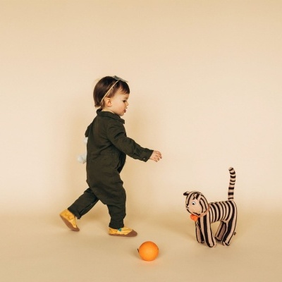 Criança com macacão verde e sapatos amarelos brinca com gato de pelúcia às riscas e laranja sobre fundo bege