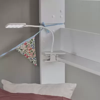 Abajur LED branco com braço flexível e base de mola preso em cabeceira de cama branca ao lado de bandeirola colorida