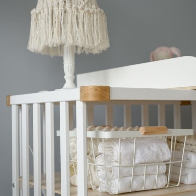 Berço branco com detalhes em madeira clara, cesta metálica com toalhas brancas e abajur trançado