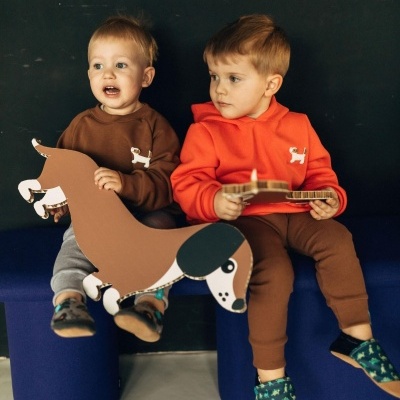 Dois meninos pequenos com camisolas castanha e laranja sentados num banco azul com brinquedos de papelão em forma de cão