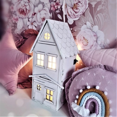 Lanterna em forma de casa branca com luz amarela entre almofadas rosas e fundo floral
