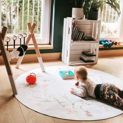 bebé deitado sobre tapete floral redondo com bolas e livros, mobiliário branco e plantas junto a janela