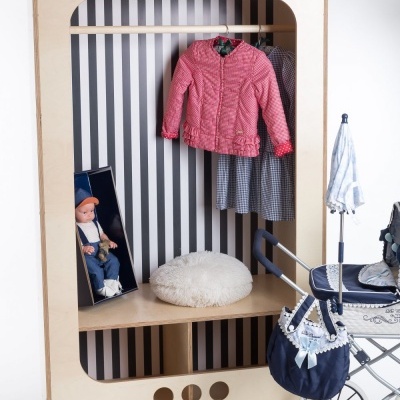Guarda-roupa infantil em madeira clara com casaco cor-de-rosa e vestido pendurados, almofada branca, boneca e carrinho de bebé azul.