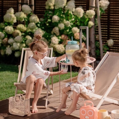 Duas crianças em espreguiçadeiras num deck de madeira com jarro de água e flores brancas ao fundo.
