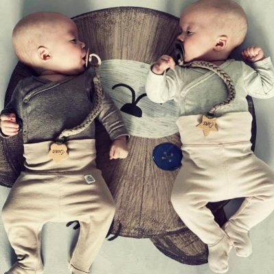 Dois bebés vestidos com macacões e babetes de algodão de cores neutras deitados num tapete com imagem de urso.