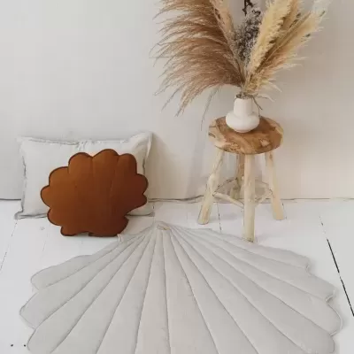 Tapete redondo em tecido bege formato concha, almofada castanha formato concha, vaso branco com flores secas em banco rústico de madeira