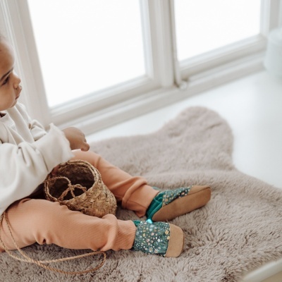 Criança sentada numa manta cinzenta com sapatinhos florais verdes e calças pêssego