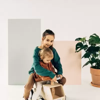 Duas crianças numa mota de madeira para crianças com painel cinza, painel rosa e planta em vaso de barro no fundo