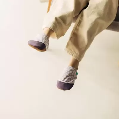 Chinelos infantis com biqueira roxa e tecido com padrão floral, usados por criança sentada numa cadeira.