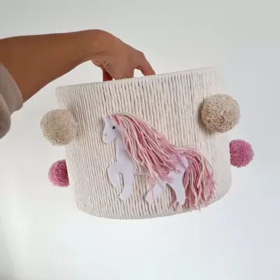 Cesto de tecido branco com pompons rosas e bege e aplique de unicórnio rosa
