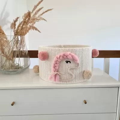Cesto decorativo branco com unicórnio rosa e pompons em cima de cómoda branca com jarro de plantas secas