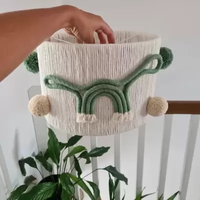 Cesto decorativo branco com arco-íris verde e pompons, em ambiente interior com plantas.