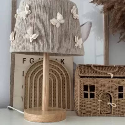 Candeeiro de mesa com asas em tecido cinzento e borboletas, base em madeira, ao lado de caixa em forma de casa de vime