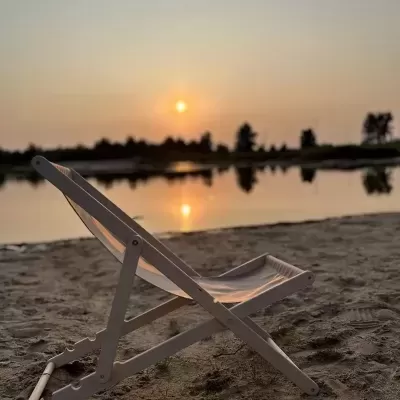 Cadeira de praia em madeira clara e tecido bege na areia junto a lago ao pôr do sol.