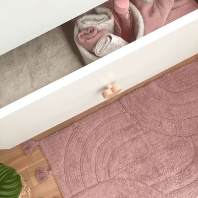 Gaveta branca aberta com toalhas rosa e brancas e embalagem rosa, sobre tapete rosa com tasséis.