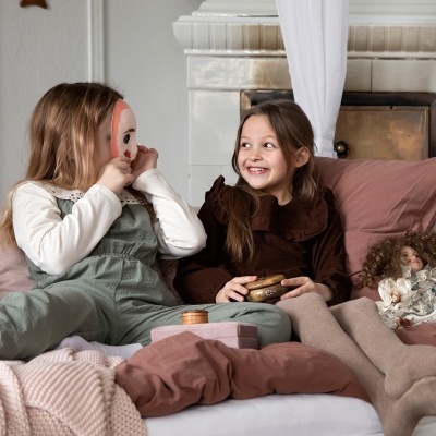 Duas meninas sentadas numa cama com roupas claras e uma máscara colorida.
