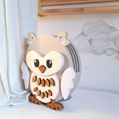 Abajur de madeira em forma de coruja com cores claras, olhos grandes e detalhes decorativos, numa mesa branca junto a cortinas e parede com nuvens desenhadas