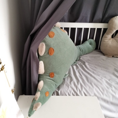 Almofada em forma de dinossauro verde e castanha sobre cama cinzenta