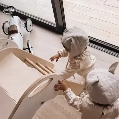 Dois bebés com roupinhas creme e capuzes, um com orelhas, brincam com brinquedo de madeira numa divisão com chão de madeira e grande janela