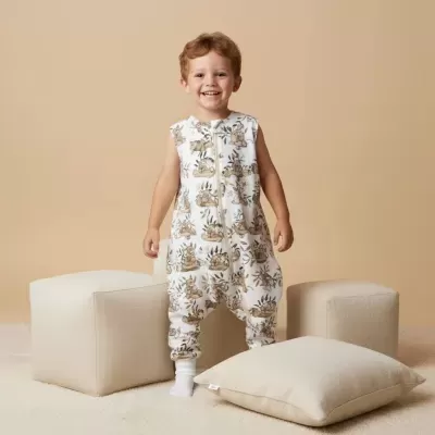 Criança a sorrir vestindo fato de dormir branco com estampado de animais em ambiente neutro com puffs e almofada bege.