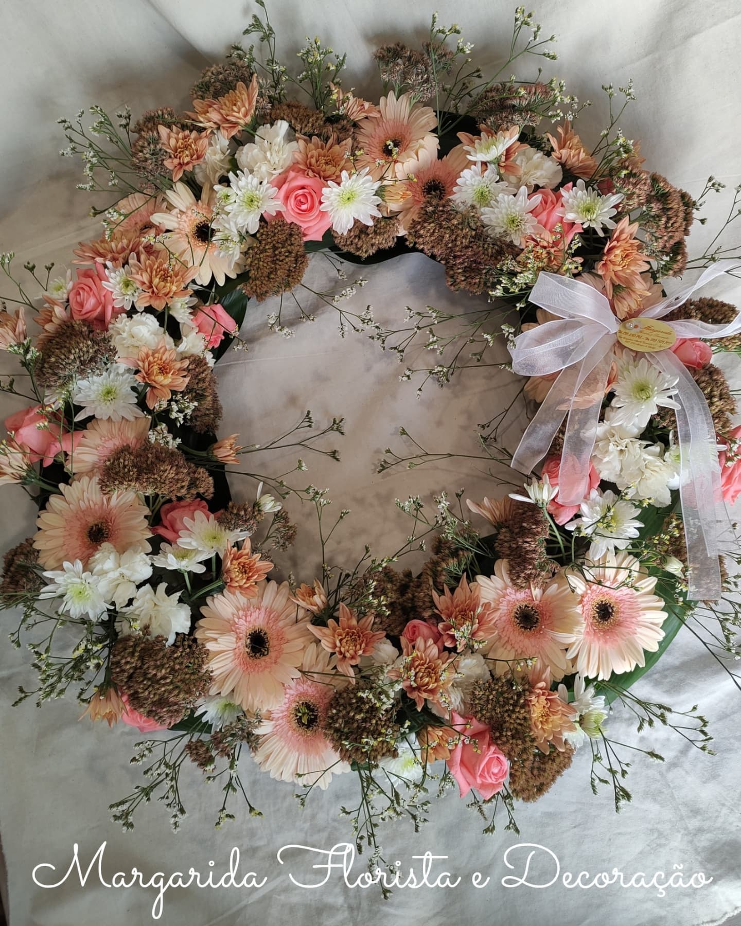 Coroa circular decorativa com flores em tons rosa, branco e castanho com laço branco.