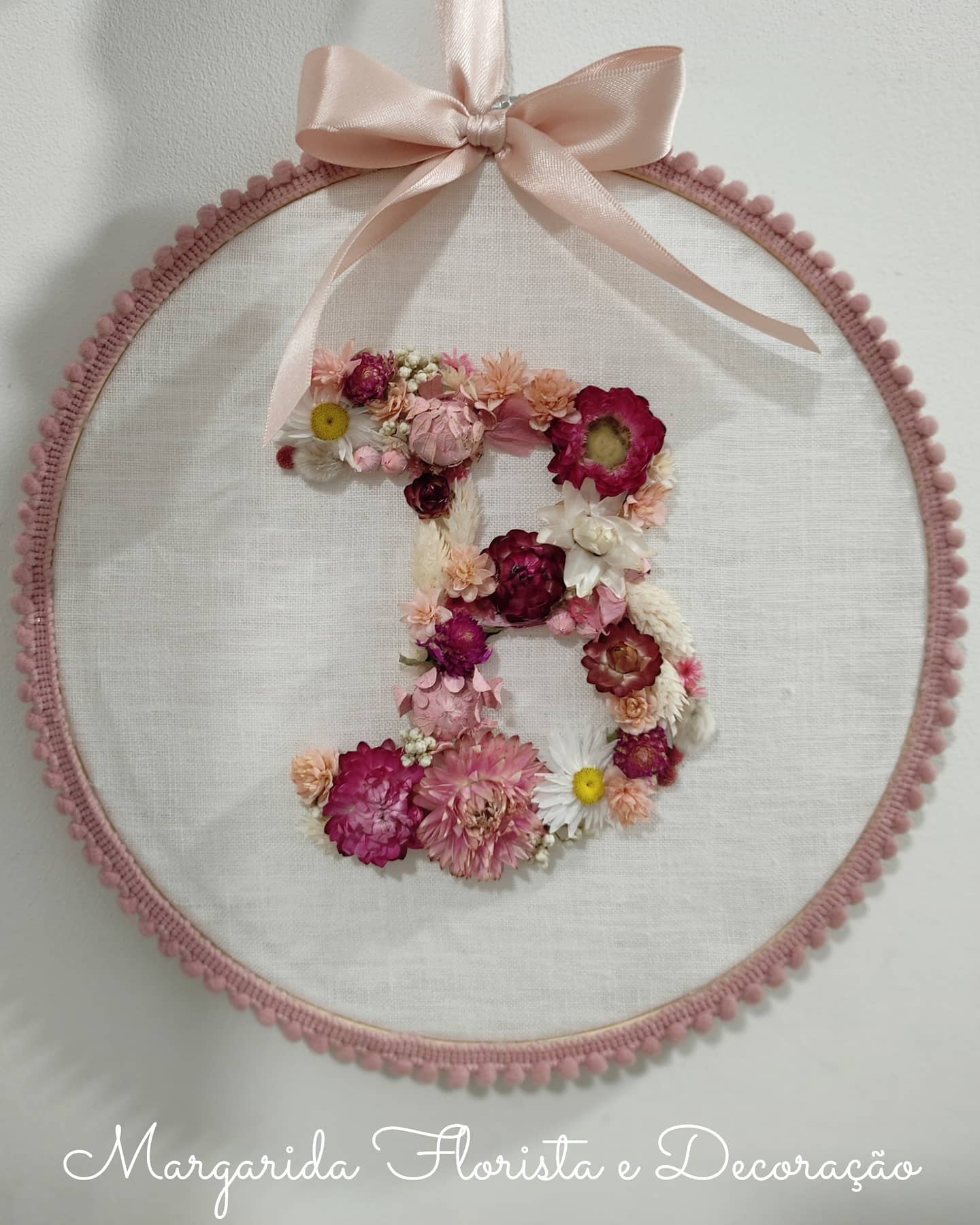 Ornamento redondo em tecido com letra B feita de flores secas e fita rosa.