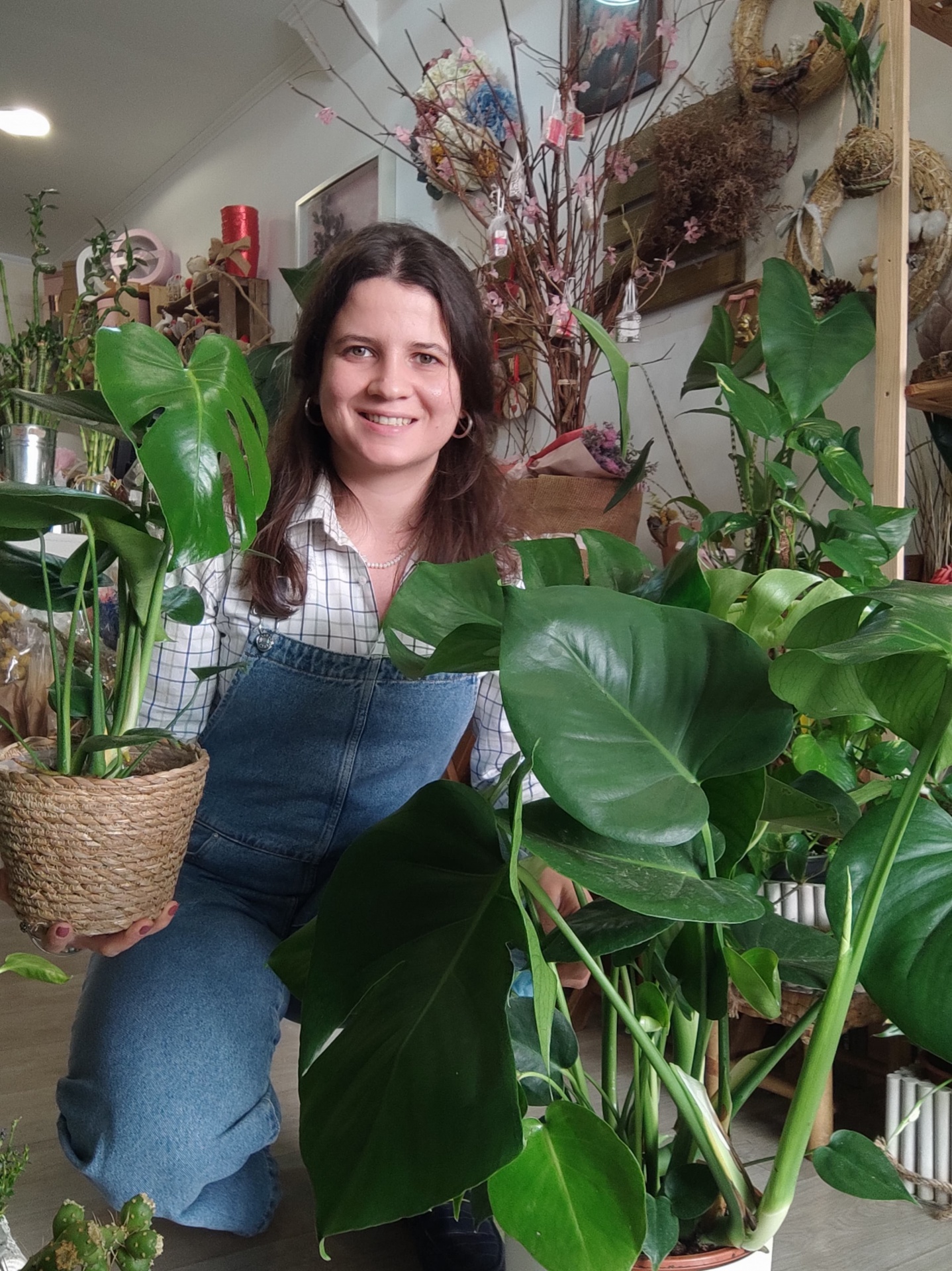 Mulher sorridente sentada entre plantas verdes em vasos, interior de casa decorado com plantas e objetos naturais.