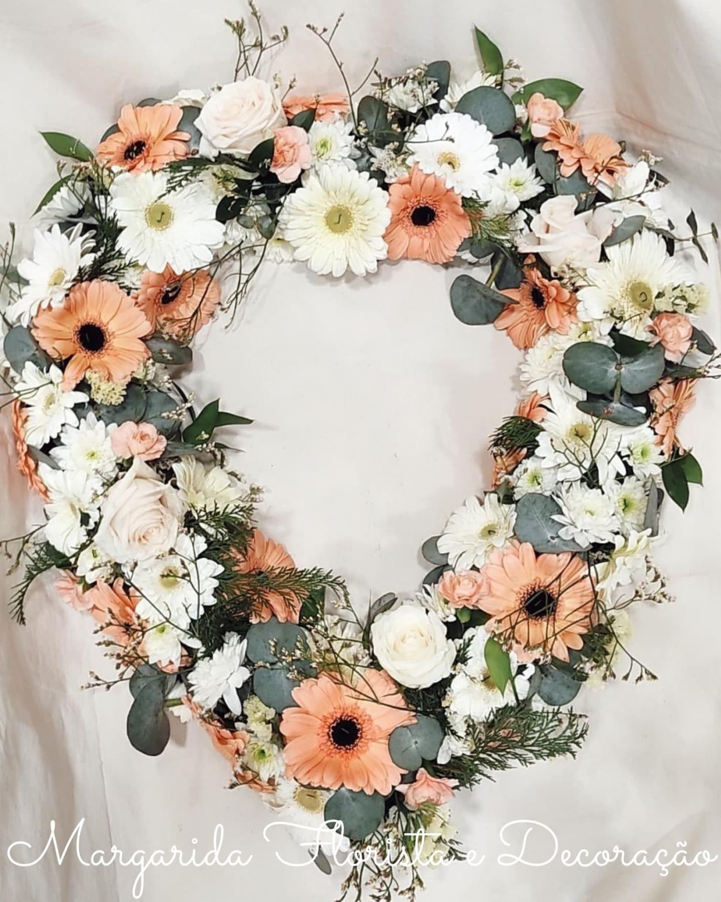 Coroa de flores artificiais branca, creme e laranja suave com folhas verdes e texto decorativo